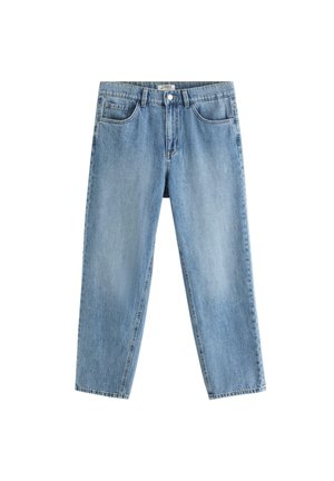 Smilende dreng står med ansigtet fremad, iført en enkel hvid t-shirt, løse blå jeans, hvide sneakers og et perlearmbånd, indendørs.