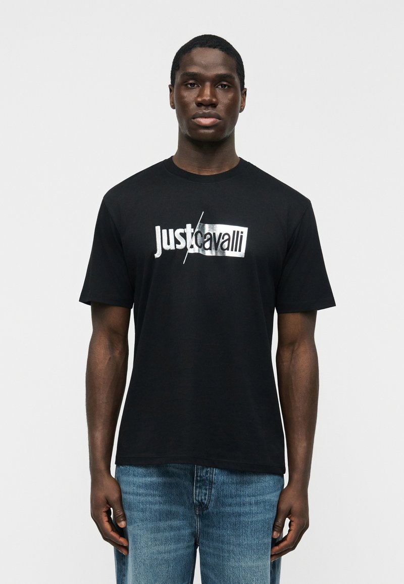 Νεαρός άνδρας που φοράει μαύρο μπλουζάκι με το λογότυπο Just Cavalli και μπλε τζιν, στέκεται μπροστά σε απλό ανοιχτόχρωμο φόντο, κοιτάζοντας προς τα εμπρός.
