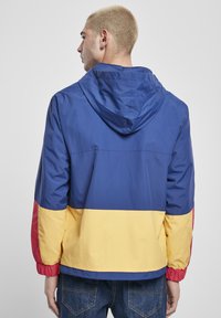 Fargeblokkert vindjakke med blå, gul og rød seksjoner. Laget av lett materiale med hette og elastiske mansjetter.