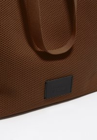 Borsa a spalla marrone con texture, realizzata in tessuto intrecciato, dotata di manici piatti e un patch con logo nero. Design semplice, spaziosa, con un aspetto robusto.