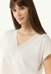 Jasno beżowa koszulka typu v-neck z krótkimi, podwiniętymi rękawami i gładką fakturą, zaprojektowana z myślą o luźnym kroju. Na tkaninie widoczny subtelny połysk.