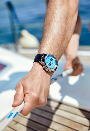 Silberner Chronograph mit einem hellblauen Zifferblatt, schwarzem Lederarmband und Druckknöpfen, getragen an einer Hand, die ein Seil gegen ein Holzdeck greift.