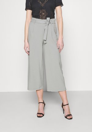Vrouw in lichtgrijze, wijde, cropped broek met een gestrikte riem, zwarte kanttop en zwarte, straps met hoge hakken sandalen.