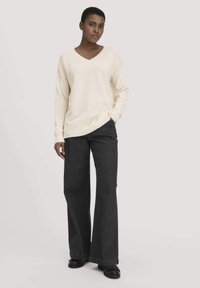 Cremfarbener V-Ausschnitt-Pullover mit langen Ärmeln, kombiniert mit dunklen weit geschnittenen Jeans. Strukturierter Strickstoff und gerippte Saumdetails. Schwarze Schuhe runden das Outfit ab.