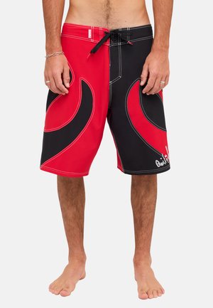Mężczyzna noszący czerwono-czarne szorty z falistymi wzorami, czarnym ściągaczem w pasie i logo Quiksilver na prawej nodze.