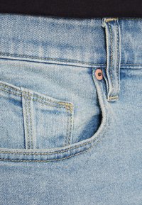 Light blue denim jeans close-up, featuring a front pocket, visible stitching, and metal button detailing. Soft texture with a faded wash.