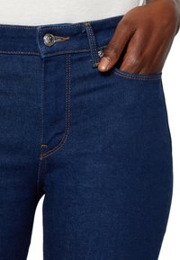 Vaqueros de denim azul oscuro con costuras naranjas, asegurados por un botón metálico, con una mano descansando dentro del bolsillo delantero.