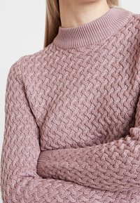Pink knitted sweater with a textured, woven pattern and a ribbed neckline, featuring long sleeves and a relaxed fit.