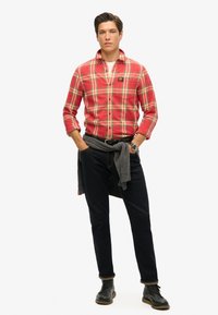 Rood geruite flanellen shirt, met knopen, gedragen over een wit T-shirt. Donkere spijkerbroek met opgerolde boorden en een grijze trui om de taille gedrapeerd. Zwarte schoenen.