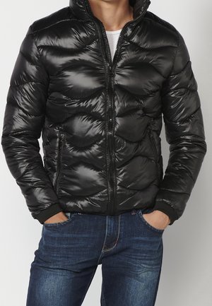 Homme portant une doudoune noire brillante sur une chemise blanche, les mains dans les poches d'un jean bleu, sur un fond uni.