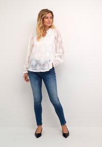 Blusa bianca in pizzo con motivo floreale, maniche lunghe e vestibilità ampia, abbinata a jeans skinny blu scuro e tacchi neri a punta.