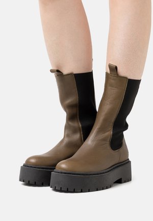 Bottes mi-mollet en cuir marron avec des panneaux élastiques noirs sur les côtés et des semelles épaisses crantées noires, portées sur des jambes nues sur un fond uni.