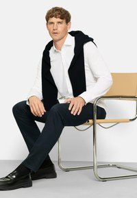 Weißes langärmeliges Hemd, dunkelblaue Hose, marineblauer Pullover über die Schultern gelegt, schwarze Schuhe, sitzt auf einem modernen Stuhl aus Metall mit einem hellbraunen Sitz.