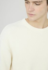 Pull en tricot couleur crème avec une texture côtelée, encolure ronde et coutures d'épaule abaissées, présentant une finition en tissu doux.