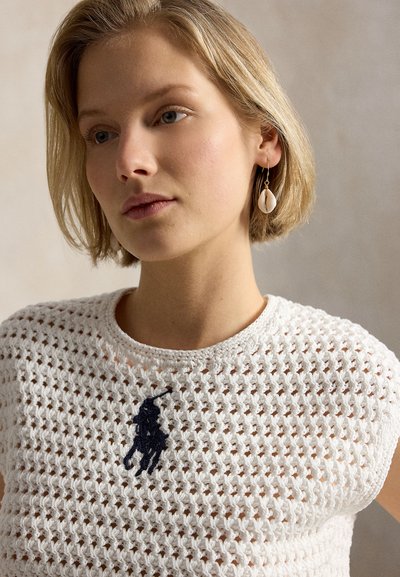 Νεαρή γυναίκα με κοντά ξανθά μαλλιά, που φοράει λευκό τοπ από πλεκτή δαντέλα με λογότυπο παίκτη πόλο σε μπλε ναυτικό χρώμα και σκουλαρίκι σε σχήμα κοχυλιού στο αριστερό αυτί.