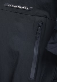Primer plano de tela de chaqueta negra con el logo de Jack & Jones y un bolsillo vertical con cremallera y pestaña texturizada.