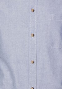 Camicia button-up azzurro chiaro realizzata in tessuto leggermente testurizzato, con bottoni color oro e una singola tasca sul petto.