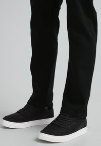 Schwarze Sportschuhe mit gerippter Textur und weißen Sohlen, kombiniert mit schwarzen Hosen. Das Design verfügt über eine Schnürung und subtile Akzente.
