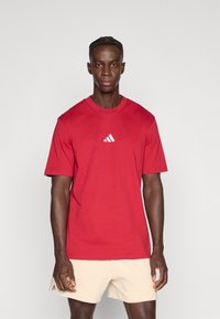 Sarkana kokvilnas T-krekls ar īsām piedurknēm, uz krūtīm baltā Adidas logotips. Audums izskatās gluds ar klasisku piegriezumu.