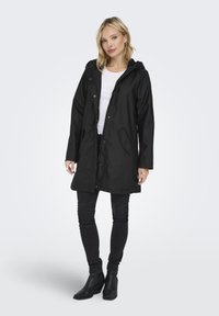 Svart vanntett frakk med hette, frontknapper, sidelommer og løs passform. Båret over en hvit strukturert topp og svarte skinny jeans.