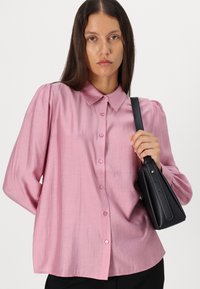 Camisa cor-de-rosa claro, de botão, com colarinho, mangas longas e volumosas, e um tecido suave; combinada com uma pequena mala preta.