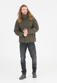 Olivenfarvet pufferjakke med høj krave, lynlåslommer og en let blank tekstur, parret med mørke jeans og grå støvler.