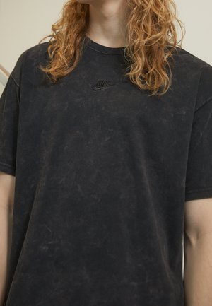 Osoba z kręconymi rudymi włosami, ubrana w wyblakły czarny t-shirt Nike z małym czarnym logo na piersi.