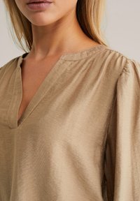 Beige Bluse mit V-Ausschnitt und plissierten Ärmeln. Der Stoff hat eine glatte Textur, mit einem subtilen Glanz und leichtem Erscheinungsbild.