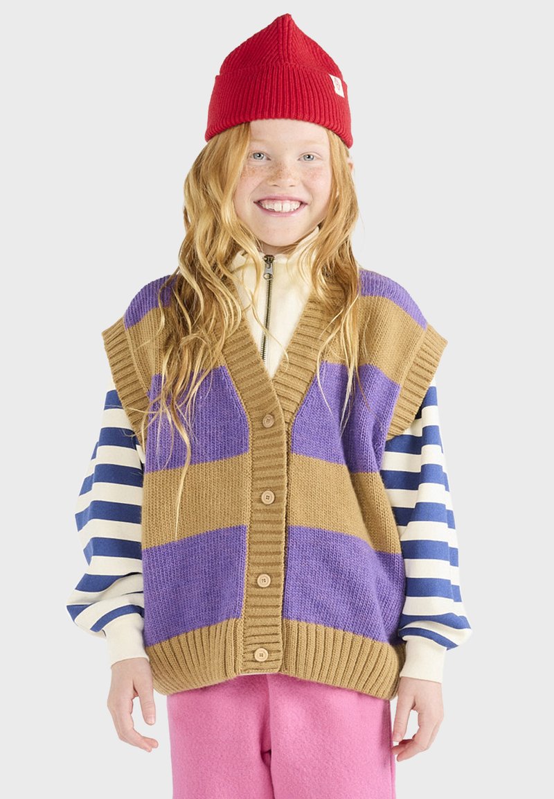 Braune und lila gestrickte Weste mit geripptem Saum und Knöpfen, getragen über einem gestreiften Longsleeve-Shirt, kombiniert mit rosa Hosen und einer roten Mütze.