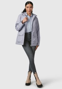 Giacca impermeabile viola chiaro con cappuccio, dotata di due tasche frontali e una texture liscia, abbinata a una camicia a righe blu e bianca e jeans neri.