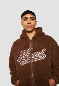 Brown fleece zip-up hoodie with a large white logo, adjustable drawstring hood, and front pockets. Soft, textured fabric.