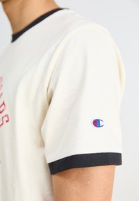 Tricou crem cu guler și manșete din bumbac ribbed de culoare albastră închisă. Are un design de text roșu și un mic logo brodat pe mâneca stângă.