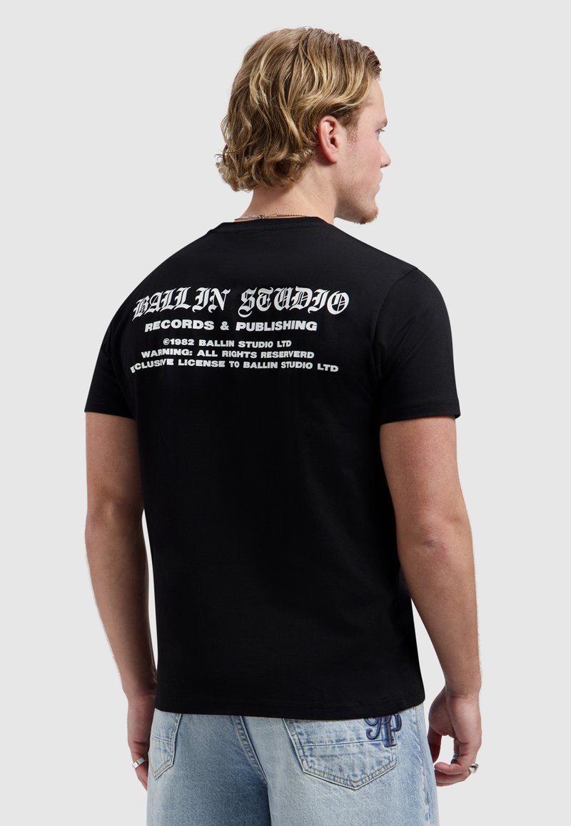 Man met blond haar die een zwart T-shirt draagt met witte tekst "Ballin Studio Records & Publishing" op de achterkant, gecombineerd met lichtblauwe spijkerbroek.
