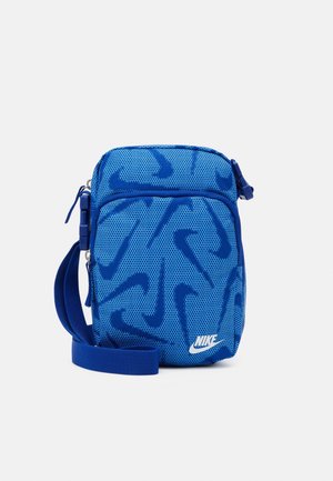 Mała niebieska torba crossbody Nike z wieloma ciemnoniebieskimi wzorami typu swoosh i regulowanym paskiem na białym tle.