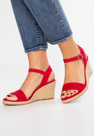 Fødder iført røde kilehælssandaler med ankelstropper og flettede espadrillestil hæle, kombineret med korte blå jeans.