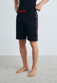 Črni bombažni šortsi z rdečim vrvico in stranskim tiskom z besedo "HUGO" v debelem tisku. Imajo elastičen pas in ravno krojenje.