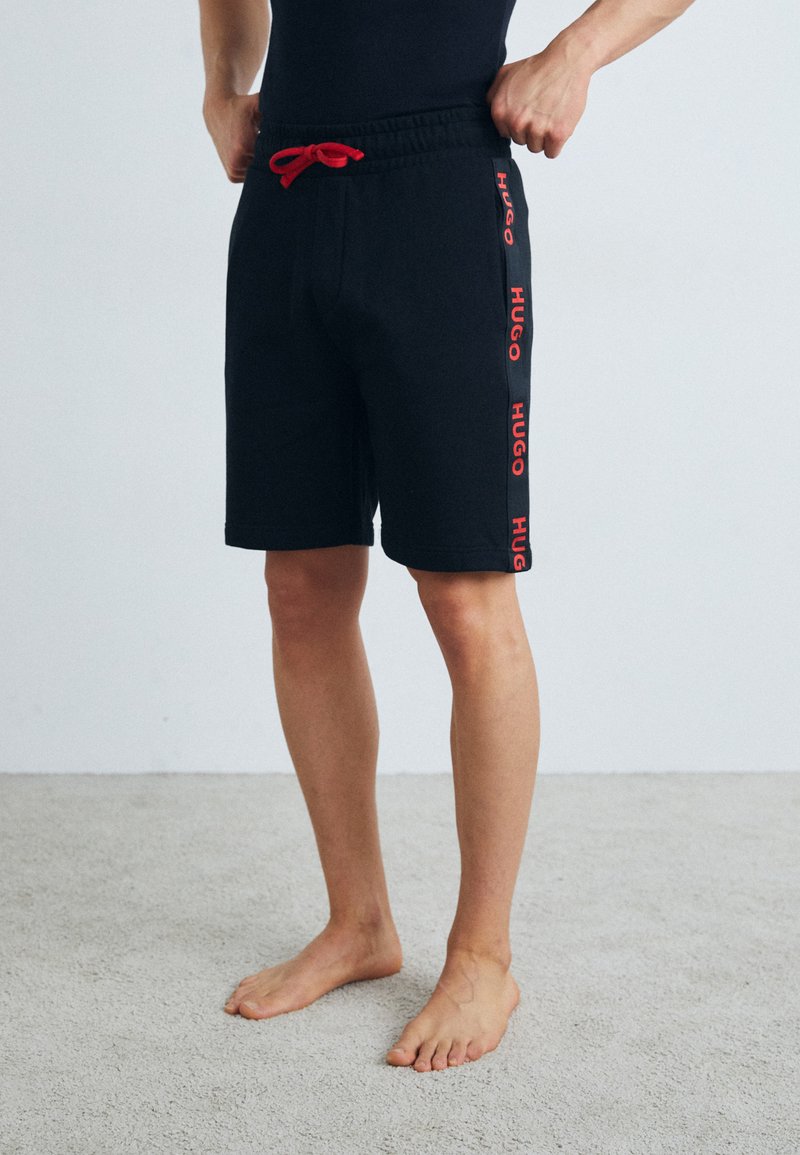 Črni bombažni šortsi z rdečim vrvico in stranskim tiskom z besedo "HUGO" v debelem tisku. Imajo elastičen pas in ravno krojenje.