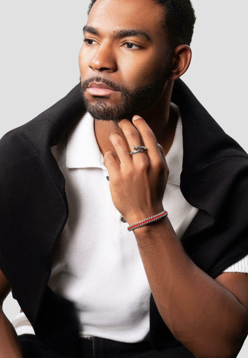 Sølvring med struktureret detaljering båret på fingre, et multicolor flettet armbånd på håndleddet, lagdelt sort sweater over en hvid polo skjorte.