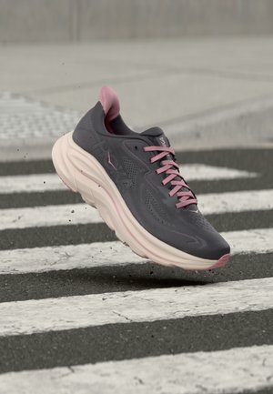 Grau-pinker Laufschuh mit dicker Sohle scheint über einen Asphaltübergang mit weißen Streifen zu springen und wirbelt kleine Schmutzpartikel auf.