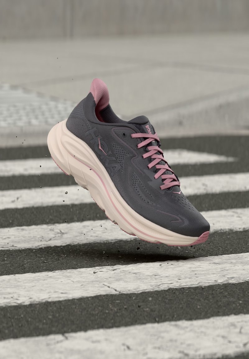 Γκρι και ροζ αθλητικό παπούτσι με χοντρή σόλα φαίνεται να πηδάει πάνω από την άσφαλτο με λευκές ρίγες, σηκώνοντας μικρά σωματίδια σκόνης.