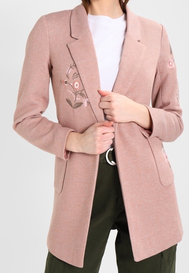 Femme portant un blazer rose poudré avec broderies florales, un haut blanc et un pantalon vert foncé, tenant les revers du blazer avec les deux mains.