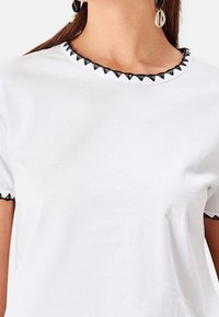 T-shirt bianco in cotone con scollo rotondo rifinito con un bordo nero a scallop, maniche corte bordate con lo stesso motivo.