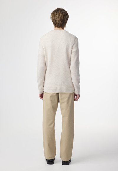 Maglione beige chiaro con una consistenza morbida e pelosa, caratterizzato da un colletto a girocollo e orlo a coste, indossato con pantaloni chiari e larghi.