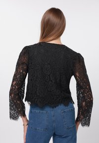 Bluză din dantelă neagră cu mâneci lungi evazate, cu modele florale și tiv în valuri. Poartă peste blugi din denim albastru.