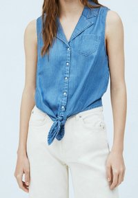 Jeansowa koszula bez rękawów z kołnierzykiem, jasnoniebieski kolor, zapięcie na guziki z przodu, jedna kieszeń na piersi, związaną na dole, w połączeniu z białymi spodniami.