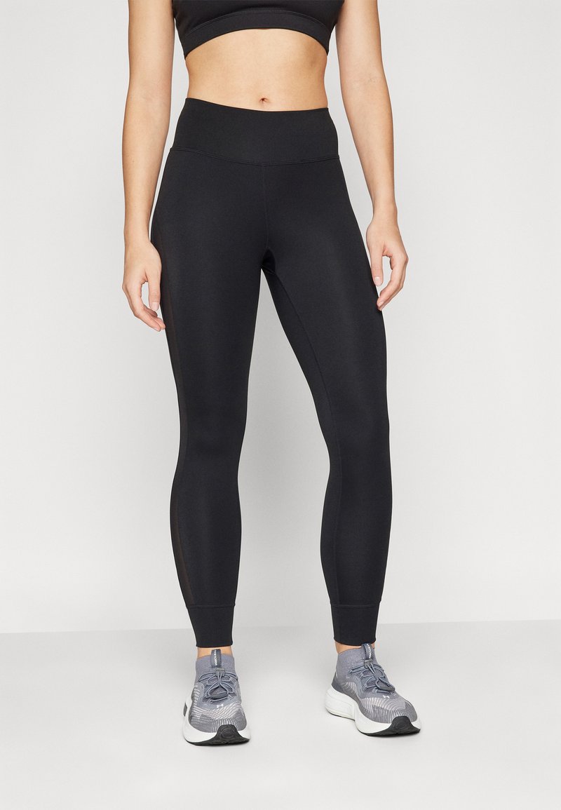 Leggings noirs à taille haute, en tissu extensible, présentant une texture lisse et une coupe à la cheville. Associés à des chaussures de sport grises.
