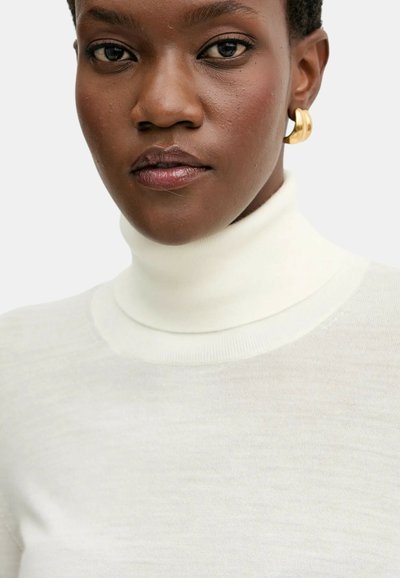 Primo piano di una persona con pelle scura che indossa un dolcevita bianco e orecchini a cerchio dorati, di fronte con un'espressione neutra.