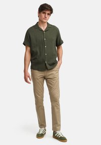 Chemise à manches courtes, couleur verte foncée, avec un tissu texturé ; associée à un pantalon kaki et des baskets vertes avec des rayures blanches.