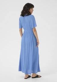 Hellblaues, knöchellanges Kleid mit kurzen Ärmeln; strukturierter Stoff, tailliert und mit fließendem Rock. Getragen mit schwarzen Riemchensandalen.