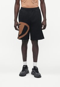 Persona de piel oscura que lleva pantalones cortos negros con un gran gráfico marrón en la pierna izquierda, calcetines blancos y zapatillas negras Diesel, de pie contra un fondo blanco.